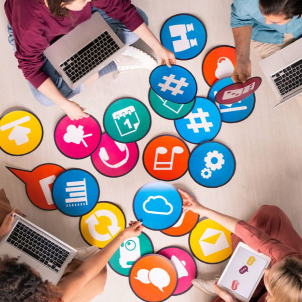 Four people sitting on floor with laptops and interacting with multiple social media platform content icons.